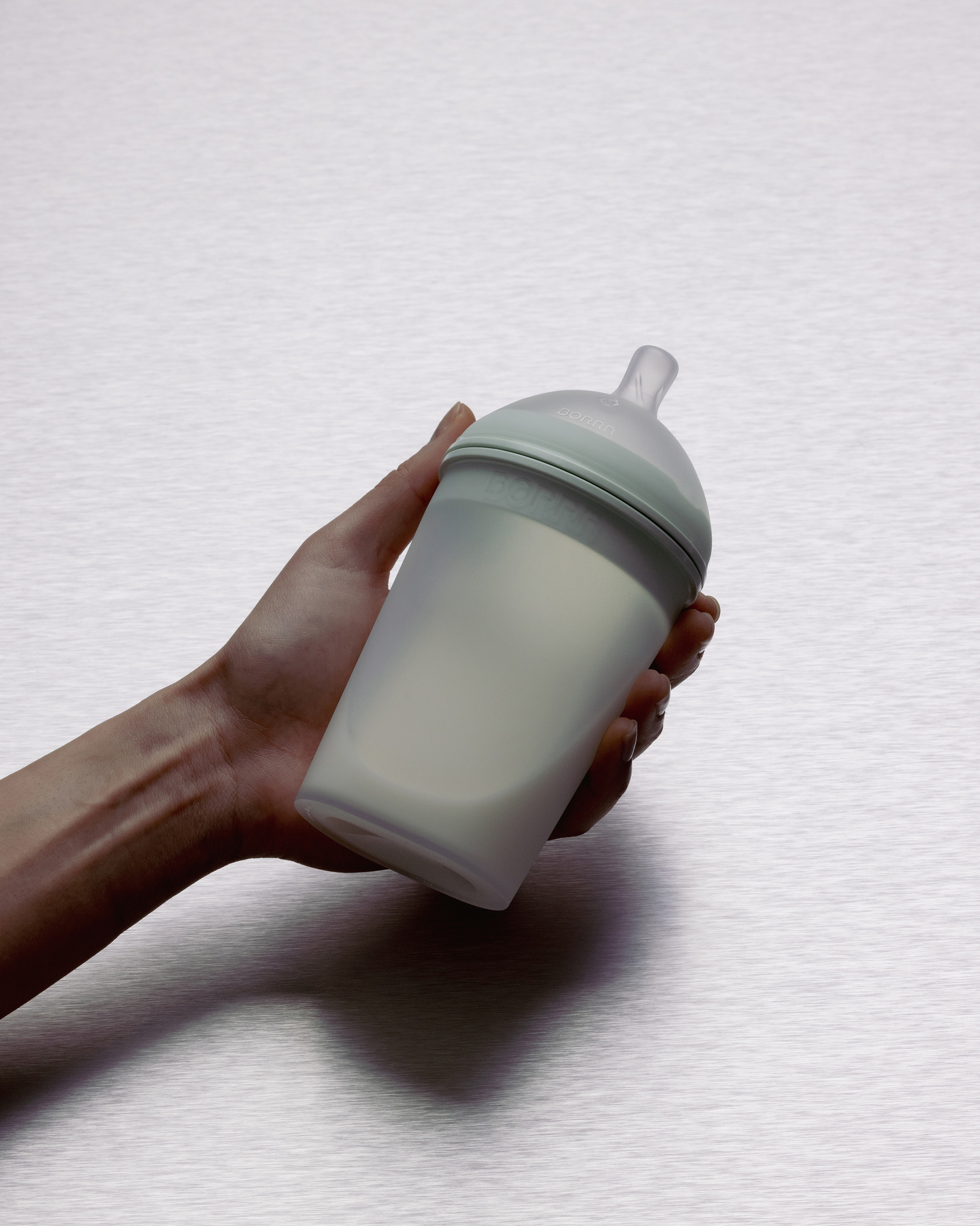 Angled close-up of hand holding translucent silicone baby feeding bottle with milk inside.